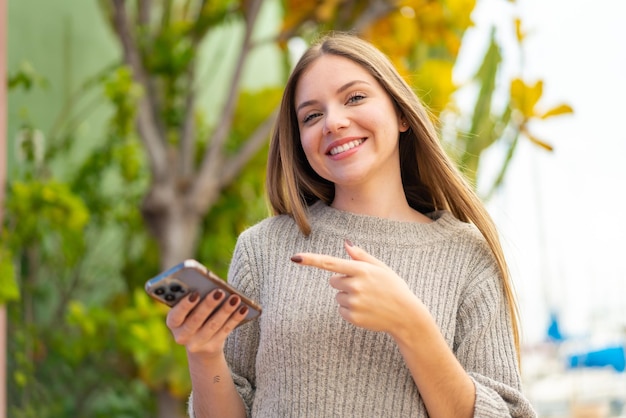 Giovane bella donna bionda che utilizza il telefono cellulare all'aperto e lo indica