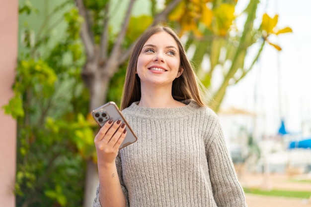 Giovane bella donna bionda che utilizza il telefono cellulare all'aperto guardando in alto mentre sorride