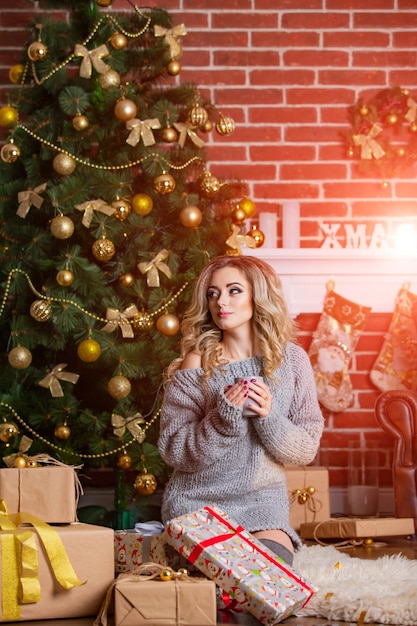 Giovane bella donna bionda è seduta sotto l'albero di Natale Decorazioni di Capodanno Sfondo muro di mattoni Ragazza in maglione grigio lavorato a maglia con regali Ritratto di una bella ragazza