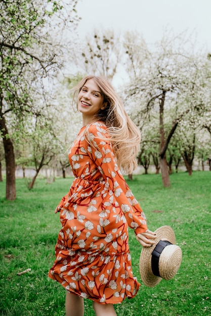Giovane bella donna bionda in giardino fiorito. Alberi primaverili in fiore Vestito arancione e cappello di paglia.