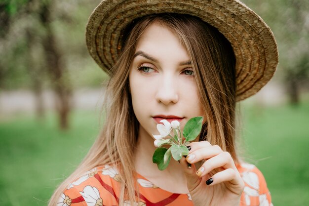 Giovane bella donna bionda in giardino fiorito. Alberi primaverili in fiore Vestito arancione e cappello di paglia.