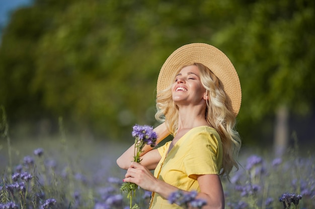 Giovane bella donna bionda in un cappello cammina attraverso un campo di fiori viola. Estate. Primavera.