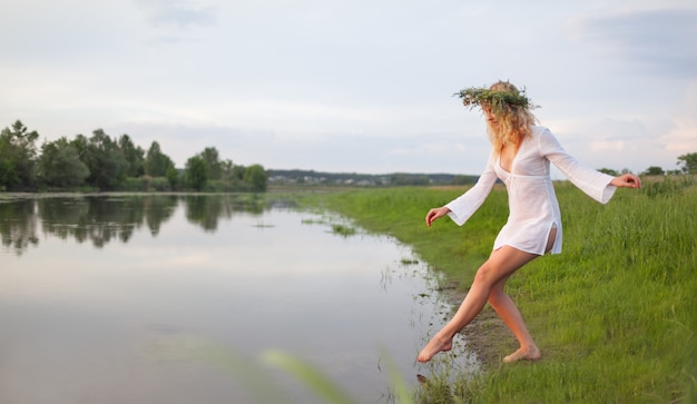 Giovane bella donna bionda sexy in mini abito bianco e corona floreale in piedi e cercando acqua