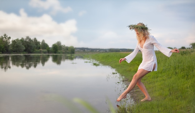 Giovane bella donna bionda sexy in mini abito bianco e corona floreale in piedi e provando l'acqua il giorno d'estate