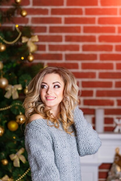 Giovane bella donna bionda vicino all'albero di Natale Decorazioni di Capodanno Sfondo muro di mattoni Preparativi per feste Ragazza in maglione grigio lavorato a maglia in uno sfondo sfocato