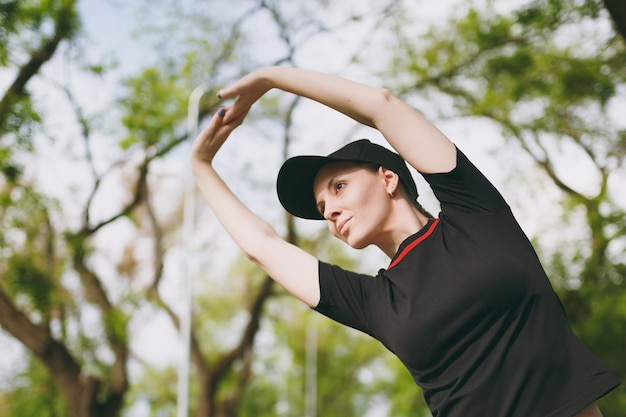 Giovane bella donna bruna atletica in uniforme nera, berretto facendo esercizi di stretching sportivo, riscaldamento prima di correre o allenarsi, in piedi nel parco cittadino all'aperto