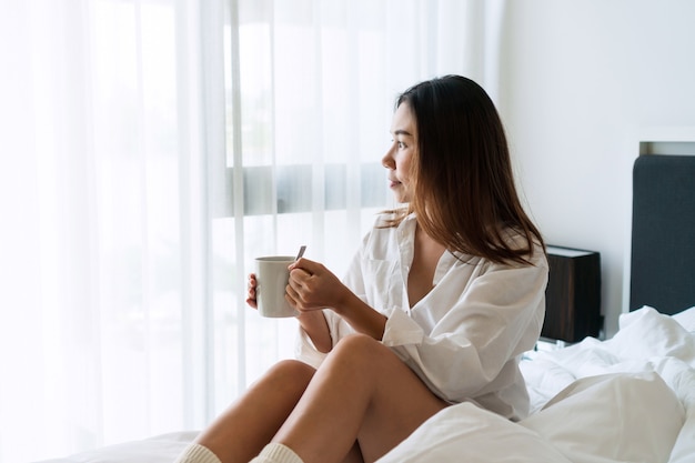 Giovane bella donna bruna capelli in pigiama camicia bianca che beve caffè mentre era seduto sul letto la mattina.