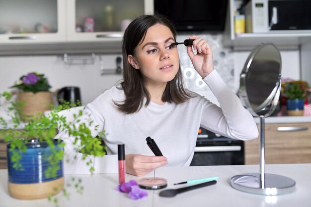 Giovane bella donna bruna che fa il trucco quotidiano per il viso con cosmetici decorativi e specchio seduto a casa in cucina, applica il mascara