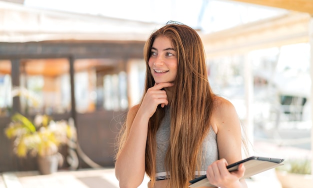 Giovane bella donna caucasica che tiene un tablet all'aperto pensando a un'idea e guardando da un lato