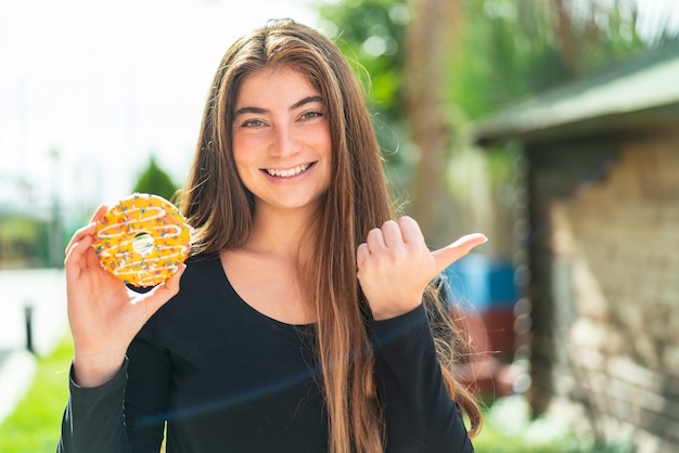 Giovane bella donna caucasica che tiene una ciambella all'aperto indicando il lato per presentare un prodotto