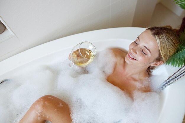 Giovane bella donna caucasica sorridente sdraiato nella vasca da bagno con schiuma e bere vino