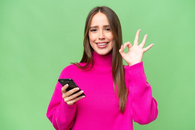 Giovane bella donna caucasica su sfondo isolato utilizzando il telefono cellulare e facendo segno OK