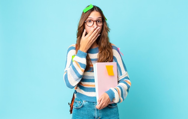 Giovane bella donna che copre la bocca con le mani con una borsa scioccata e tiene in mano dei libri