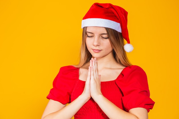 Giovane bella donna che indossa il cappello di Babbo Natale su sfondo giallo isolato, pregando con le mani insieme chiedendo perdono, sorridendo con fiducia.
