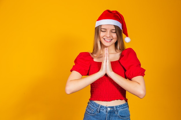 Giovane bella donna che indossa il cappello di Babbo Natale su sfondo giallo isolato, pregando con le mani insieme chiedendo perdono, sorridendo con fiducia.