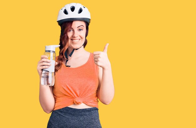 Giovane bella donna che indossa il casco da bici e tiene la bottiglia d'acqua sorridendo felice e positivo, pollice in su facendo eccellente e segno di approvazione