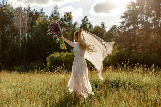 Giovane bella donna che indossa un abito bianco con in mano fiori e balla sul prato Ragazza che gioisce della natura e della libertà Bellezza naturale Movimento di danza Salute mentale Sognare senza stress Tramonto