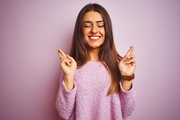 Giovane bella donna che indossa un maglione casual in piedi su sfondo rosa isolato gesticolare dito incrociato sorridendo con speranza e occhi chiusi Fortuna e concetto superstizioso