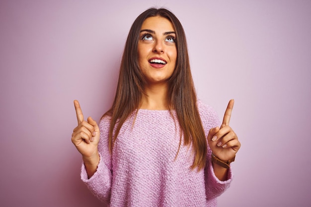 Giovane bella donna che indossa un maglione casual in piedi su sfondo rosa isolato stupito e sorpreso guardando in alto e puntando con le dita e le braccia sollevate