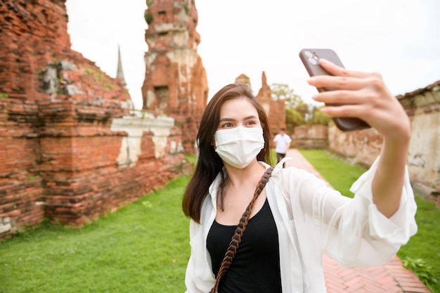 Giovane bella donna che indossa una maschera protettiva che viaggia e scatta foto al parco storico tailandese