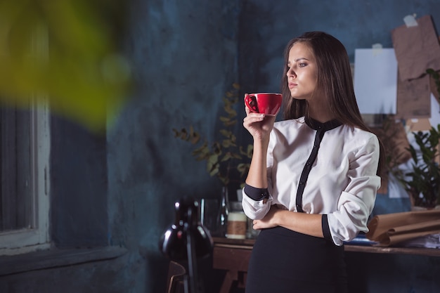 Giovane bella donna che lavora con una tazza di caffè e notebook presso l'ufficio loft