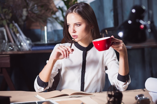 Giovane bella donna che lavora con una tazza di caffè e un taccuino in ufficio loft