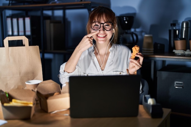 Giovane bella donna che lavora utilizzando il computer portatile e mangia cibo di consegna che punta con il dito della mano al viso e al naso, sorridendo allegro. concetto di bellezza