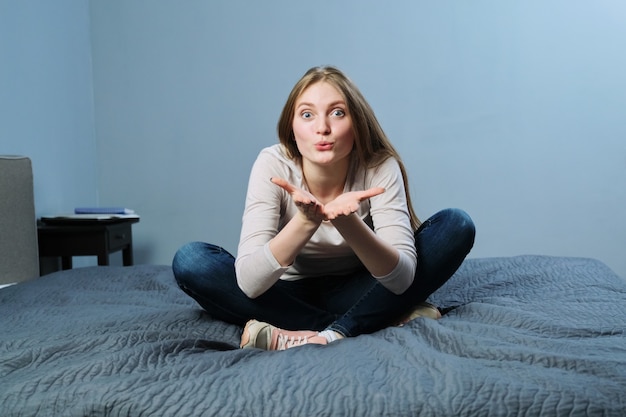 Giovane bella donna che mostra le labbra con un bacio, la donna guarda la telecamera, ti fa un gesto, seduta a casa a letto. Emozione amore, felicità, gioia, concetto di giovani