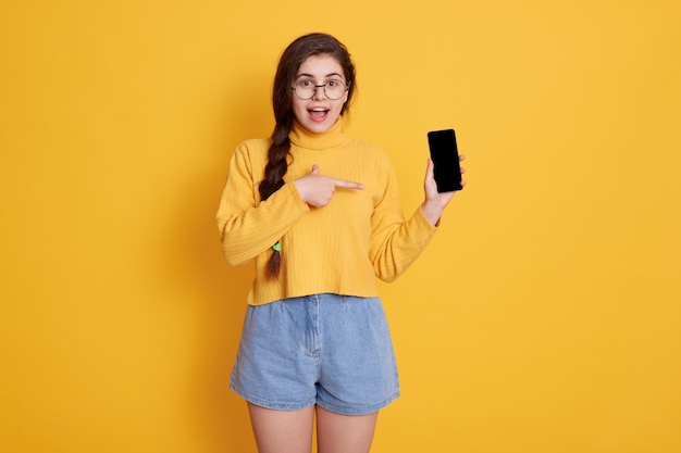 Giovane bella donna che mostra lo schermo vuoto dello smart phone con il dito indice isolato sopra la parete gialla, mantenendo la bocca aperta, indossando abiti eleganti.