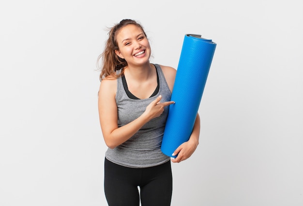 Giovane bella donna che sembra eccitata e sorpresa indicando il lato e tenendo in mano un tappetino da yoga