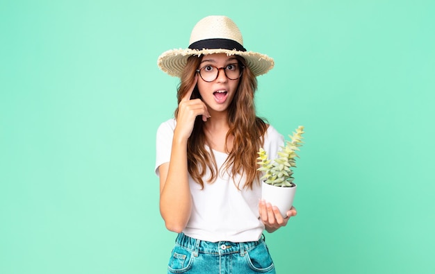 Giovane bella donna che sembra sorpresa, realizzando un nuovo pensiero, idea o concetto con un cappello di paglia e tenendo in mano un cactus