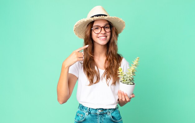 Giovane bella donna che sorride con sicurezza indicando il proprio ampio sorriso con un cappello di paglia e tenendo in mano un cactus