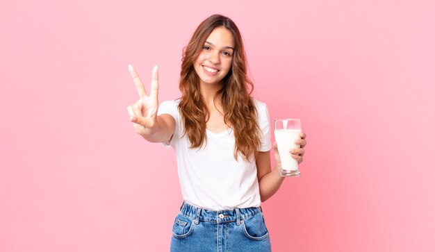 Giovane bella donna che sorride e sembra felice, gesticola vittoria o pace e tiene in mano un bicchiere di latte