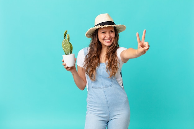 Giovane bella donna che sorride e sembra felice, gesticola vittoria o pace e tiene in mano un cactus