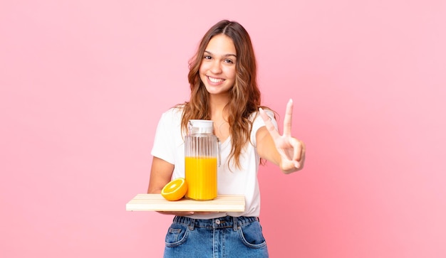 Giovane bella donna che sorride e sembra felice, gesticola vittoria o pace e tiene in mano un vassoio con un succo d'arancia