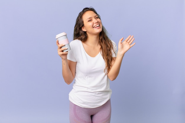 giovane bella donna che sorride felice, agitando la mano, accogliendoti e salutandoti e tenendo in mano un caffè