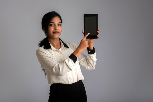 Giovane bella donna che tiene e che mostra smartphone schermo vuoto o cellulare o tablet su un muro grigio.