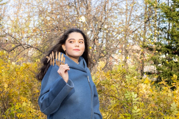 Giovane bella donna che tiene le foglie gialle in mano. Paesaggio autunnale in una giornata di sole. Una ragazza allegra con i capelli lunghi in un cappotto sorride in una calda giornata autunnale sullo sfondo degli alberi del parco