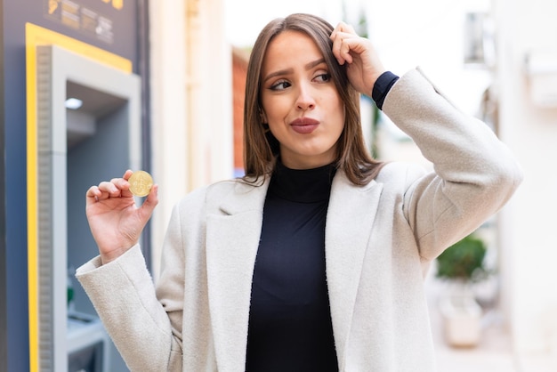 Giovane bella donna che tiene un Bitcoin all'aperto con dubbi e con espressione del viso confusa