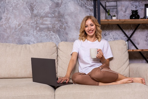 Giovane bella donna che utilizza computer portatile sorridendo felice guardando e navigando o facendo acquisti online su Internet a casa. Soggiorno moderno. Espressione del viso allegra e soddisfatta