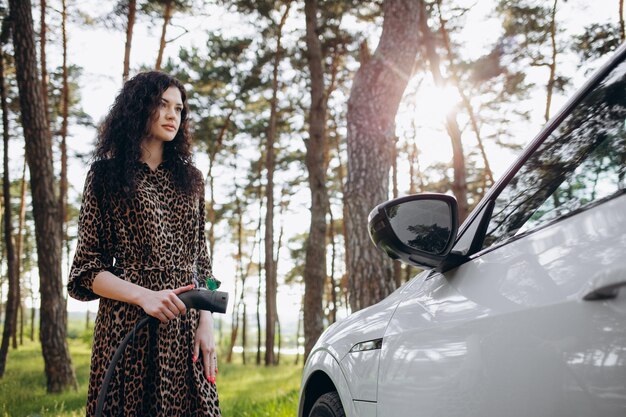 Giovane bella donna che viaggia in auto elettrica che si ferma alla stazione di ricarica in piedi collegando il cavo navigando in internet sullo smartphone sorridendo gioioso mentre si carica