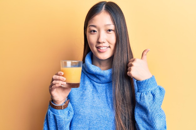 Giovane bella donna cinese che beve bicchiere di succo d'arancia sorridendo felice e positivo, pollice in su facendo eccellente e segno di approvazione