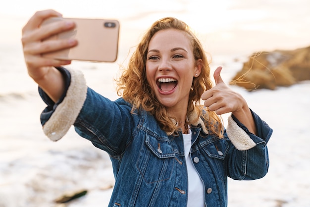 giovane bella donna con capelli biondi ricci utilizzando smartphone mentre si cammina in riva al mare