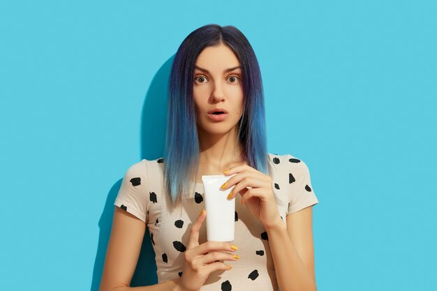 Giovane bella donna con capelli blu che tiene in mano un flacone cosmetico bianco