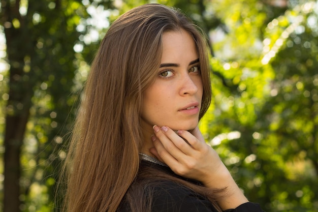 Giovane bella donna con capelli lunghi castani in camicetta nera e collana d'argento in piedi sullo sfondo di alberi verdi