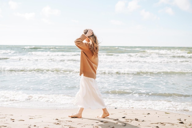 Giovane bella donna con capelli lunghi in maglione beige che si gode la vita sulla spiaggia del mare al tramonto