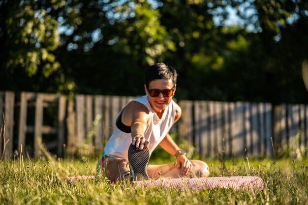 Giovane bella donna con capelli neri corti che si allungano su un tappetino da yoga su un prato in un parco, riscaldarsi prima dell'allenamento