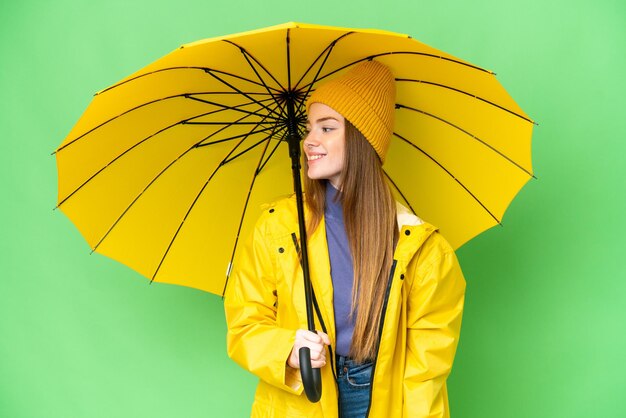Giovane bella donna con cappotto antipioggia e ombrello su sfondo chiave cromatica isolato guardando lato