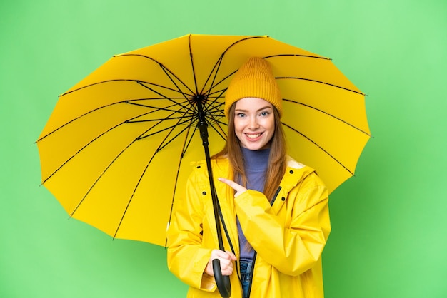 Giovane bella donna con cappotto antipioggia e ombrello su sfondo chroma key isolato che punta al lato per presentare un prodotto