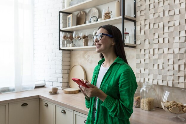 Giovane bella donna con gli occhiali e camicia verde vicino alla finestra di casa in cucina usando un rosso